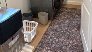A laundry room with stacked washers and dryers, a washing machine, a dryer, a trash bin, a laundry basket, and a small table with a power outlet.