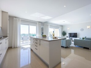 Eine moderne Küche mit Kochinsel, großen Fenstern und einem Wohnbereich mit Sofa und Fernseher.