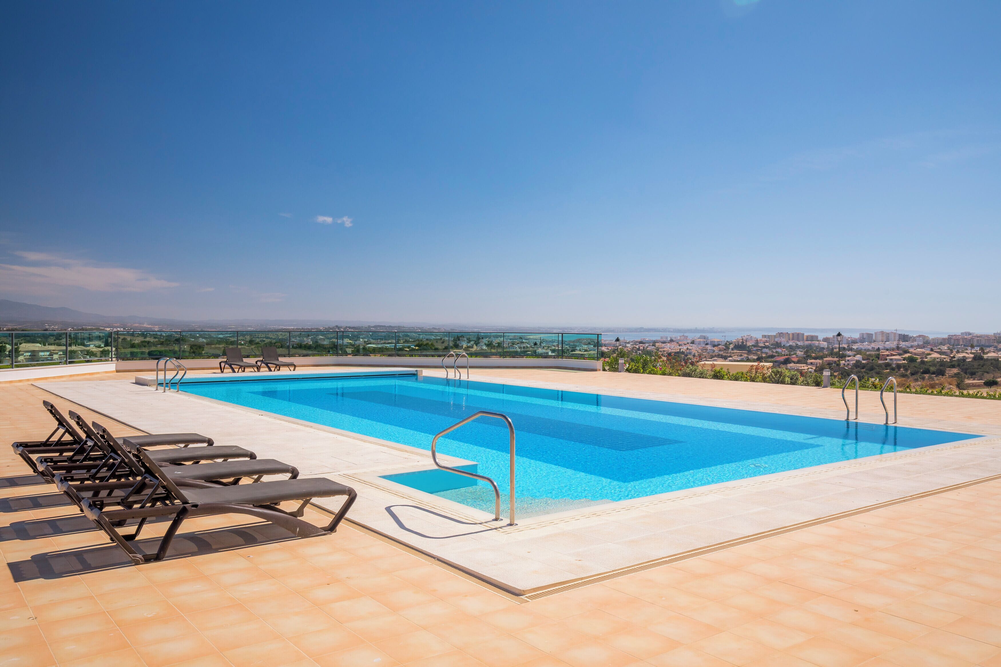 A rooftop swimming pool with clear blue water, surrounded by lounge chairs and a city view in the background.