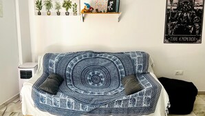 Een kamer met een blauw-witte bank met een patroon, een klein rekje met planten en decoratieve items, en een ingelijst schilderij aan de muur.