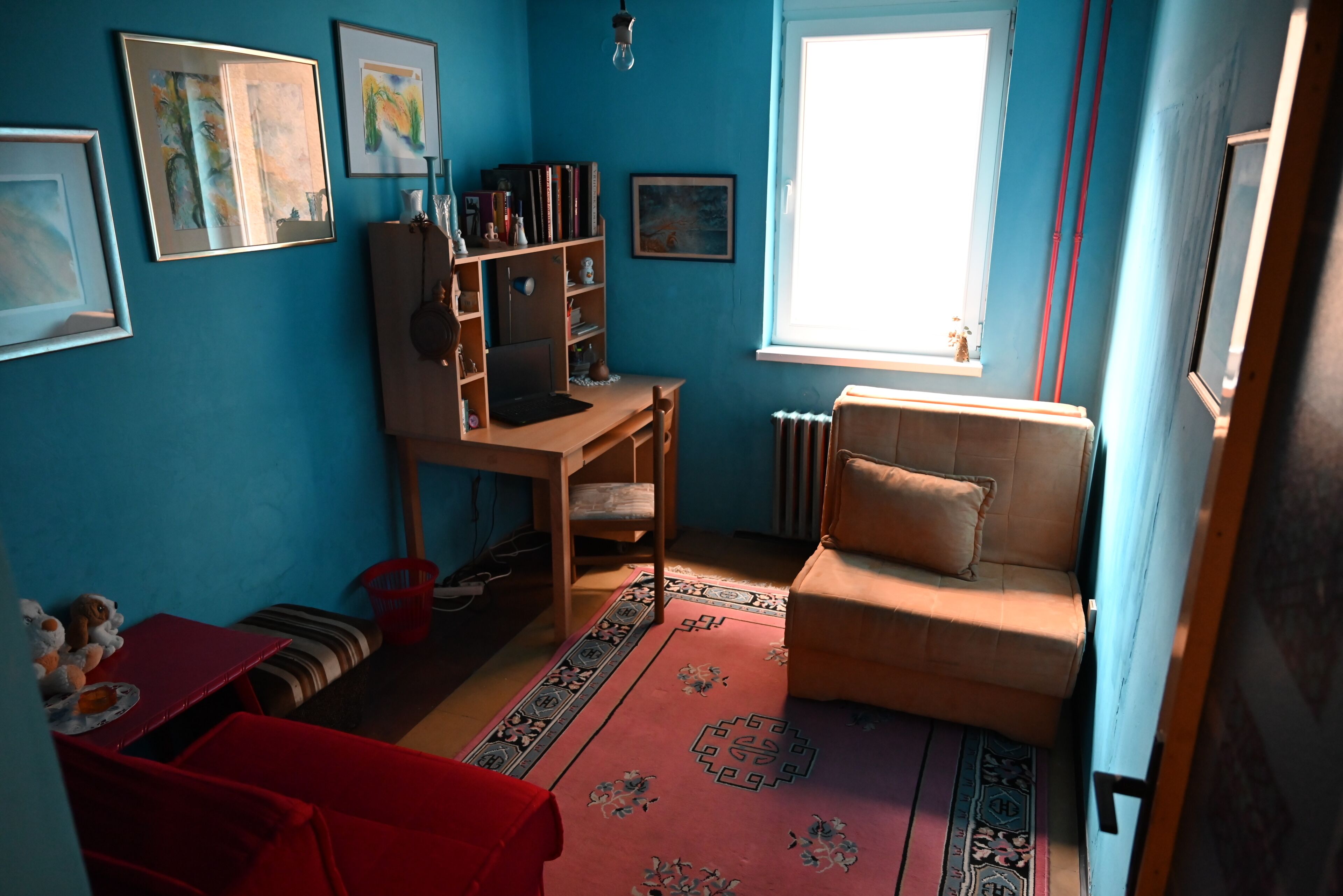 A room with a red sofa, a wooden desk, a bookshelf, and a window with a view.