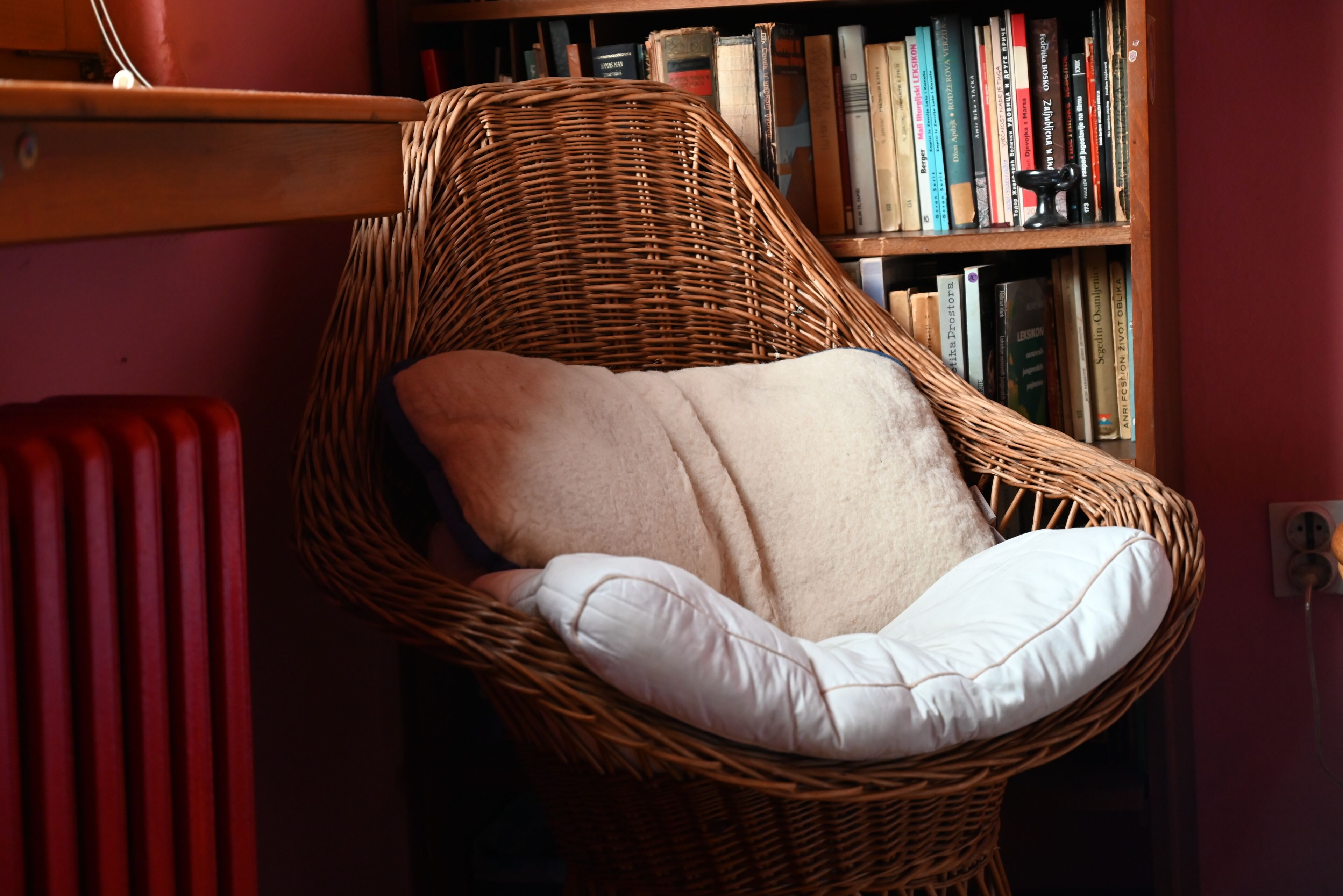 Een rieten stoel met een witte kussens op een kamer, met een boekenplank op de achtergrond.