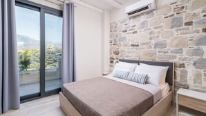 A bedroom with a stone wall, a bed with pillows, a nightstand, and a window with curtains. - Elmara - Beachside Lux Villa 2 With Private Pool (Faistos)