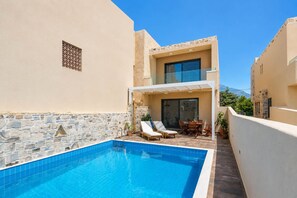 A swimming pool with lounge chairs, a stone wall, and a modern building with a glass door. - Elmara - Beachside Lux Villa 2 With Private Pool (Faistos)