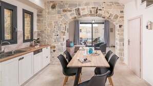 A modern kitchen with stone walls, a dining area with a wooden table and chairs, and a view through an archway to a balcony. - Elmara - Beachside Lux Villa 2 With Private Pool (Faistos)