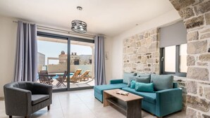 A living room with a sofa, armchair, and coffee table, a stone wall, and a view through a glass door. - Elmara Lux Seaside Villa 1 Private Pool in Crete (Faistos)