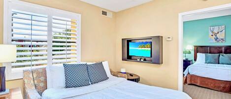 A hotel room with a bed, a TV mounted on the wall, a window with blinds, and a door leading to another room.