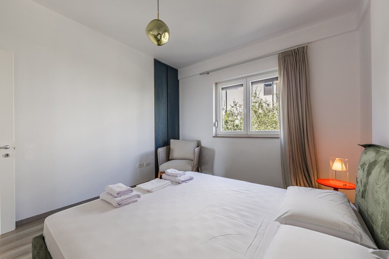 A hotel room with a bed, a chair, a window with curtains, and a bedside table with a lamp.