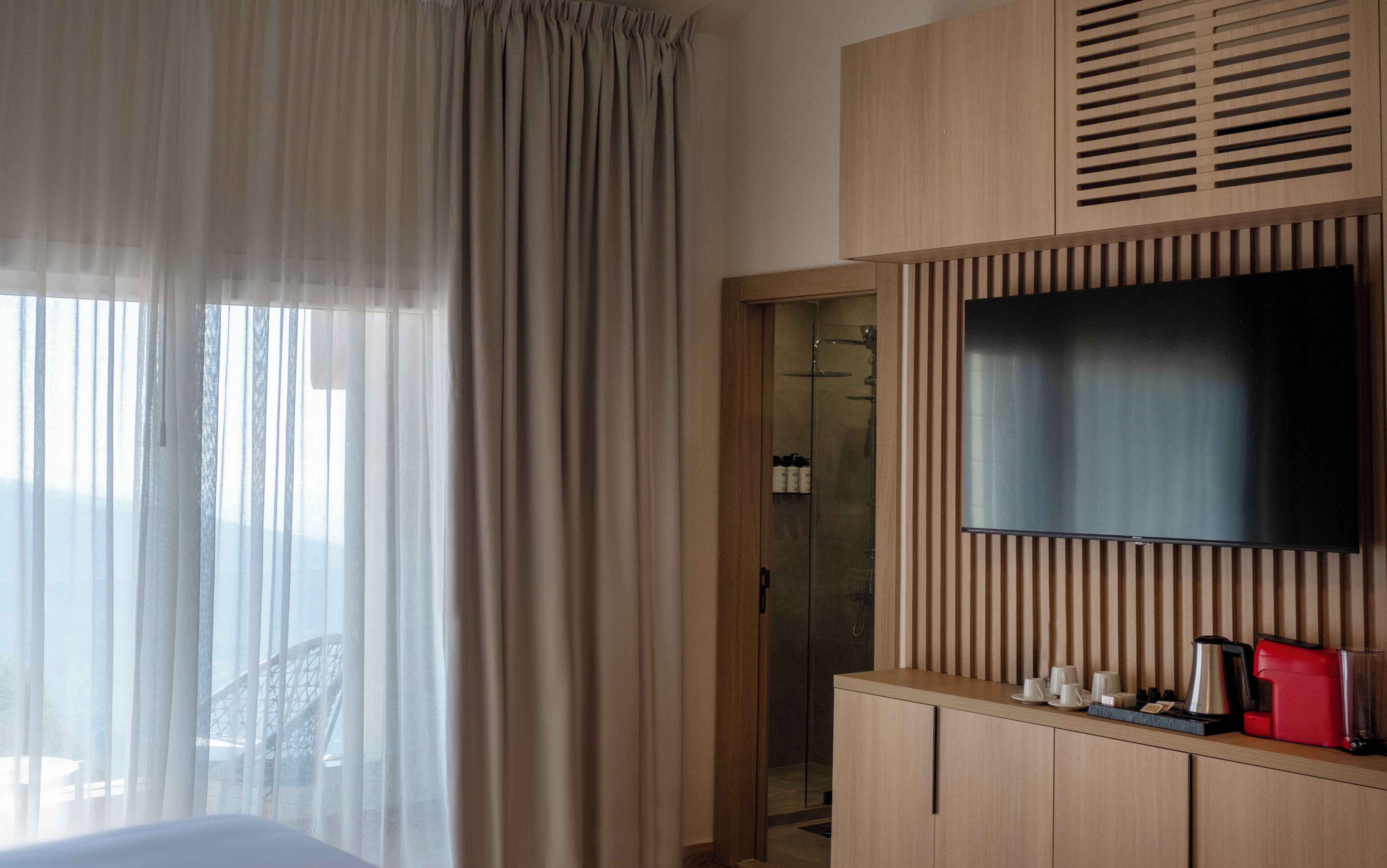 A hotel room with a bed, a TV mounted on a wooden panel, a glass shower door, and a window with curtains.