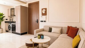 Una sala moderna con un sofá, una mesita de centro y una mesita auxiliar pequeña. En la cocina contigua hay una lavadora y un microondas.