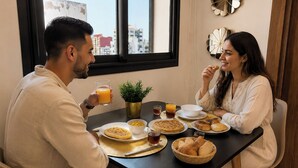 Una mesa de comedor con alimentos para el desayuno, una planta en maceta y vistas de edificios a través de una ventana.