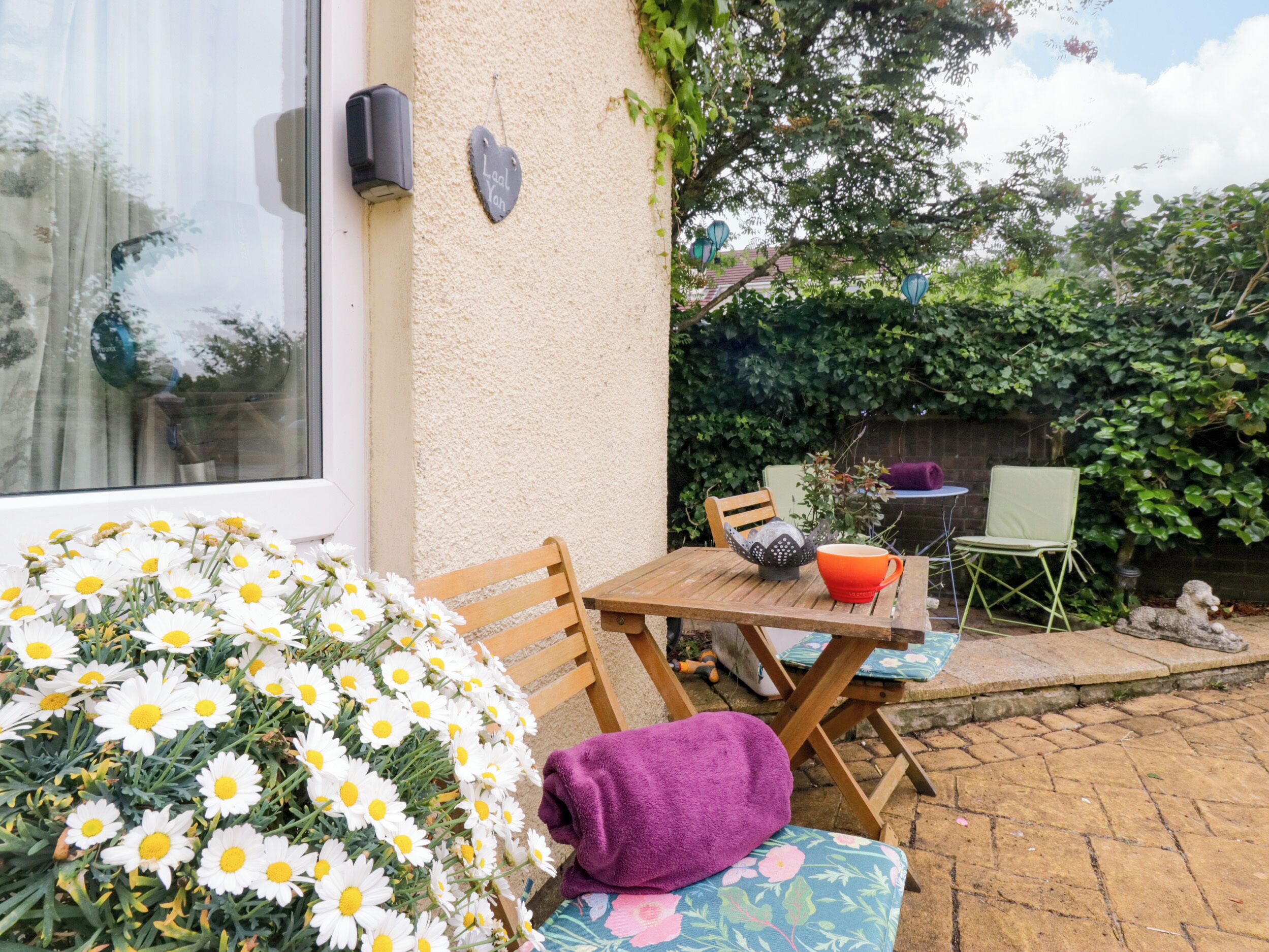 Een terras met een houten tafel, stoelen, een paarse handdoek en een bloemstuk.