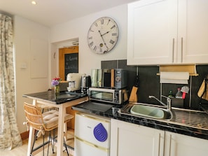 Una cocina compacta con un reloj, un tostador, un microondas, un lavabo y un pequeño comedor con una silla.