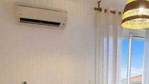 Een woonkamer met een bank, twee kussens, een houten salontafel, een airconditioner en uitzicht door een raam.