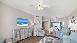 Una sala de estar con ventilador de techo, televisor, comedor con sillas y alfombra estampada.