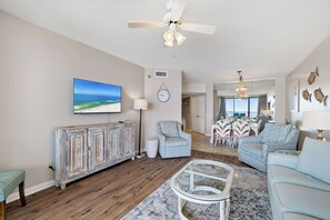 A living room with a ceiling fan, a television, a dining area with chairs, and a patterned rug. - White Caps 505 by Liquid Life (Orange Beach)