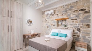 A bedroom with a stone wall, a bed with a beige bedspread, a desk with a chair, and a wardrobe. - Elmara Crete Beachside Villa 3 With Private Pool (Faistos)
