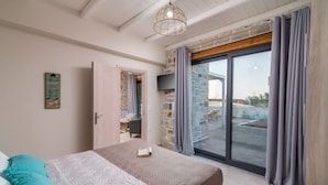 A bedroom with a bed, a TV, a window with curtains, and a stone wall. - Elmara Crete Beachside Villa 3 With Private Pool (Faistos)