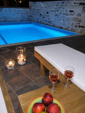 Ein Poolbereich mit Steinmauer, einer hölzernen Bank, zwei Kerzen, zwei Gläser Roséwein und einem Teller mit Äpfeln.