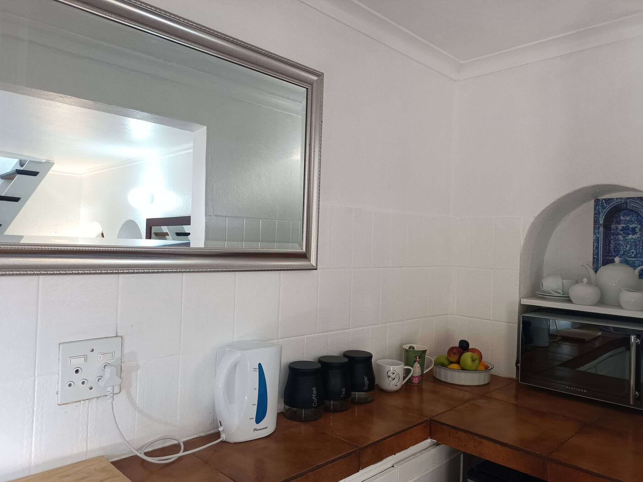 A kitchen with white tiles, a large mirror, a coffee maker, a kettle, a microwave, and a fruit bowl.