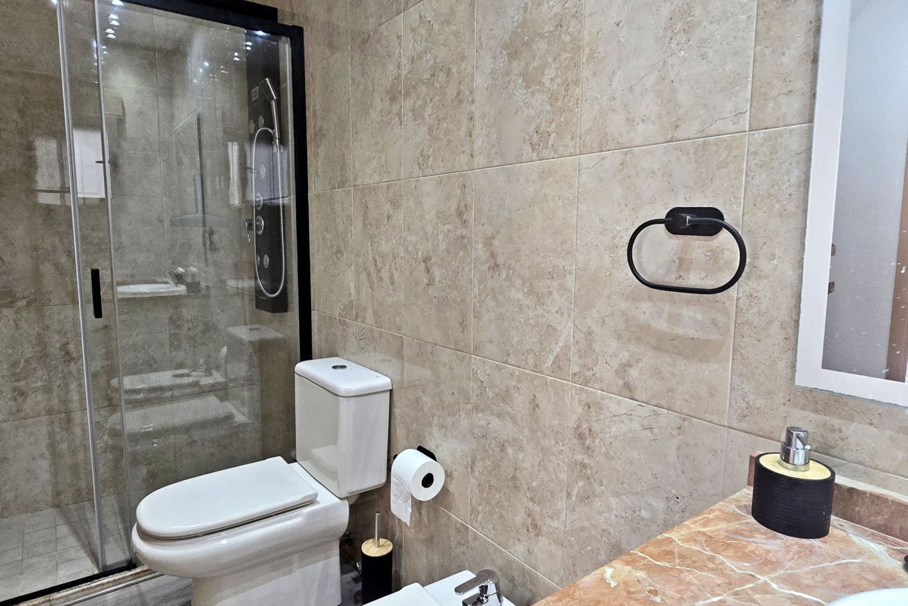Une salle de bain comprenant des toilettes, une douche en verre, un mur recouvert de carreaux de marbre, un porte-rouleau de papier toilette et un porte-serviettes.