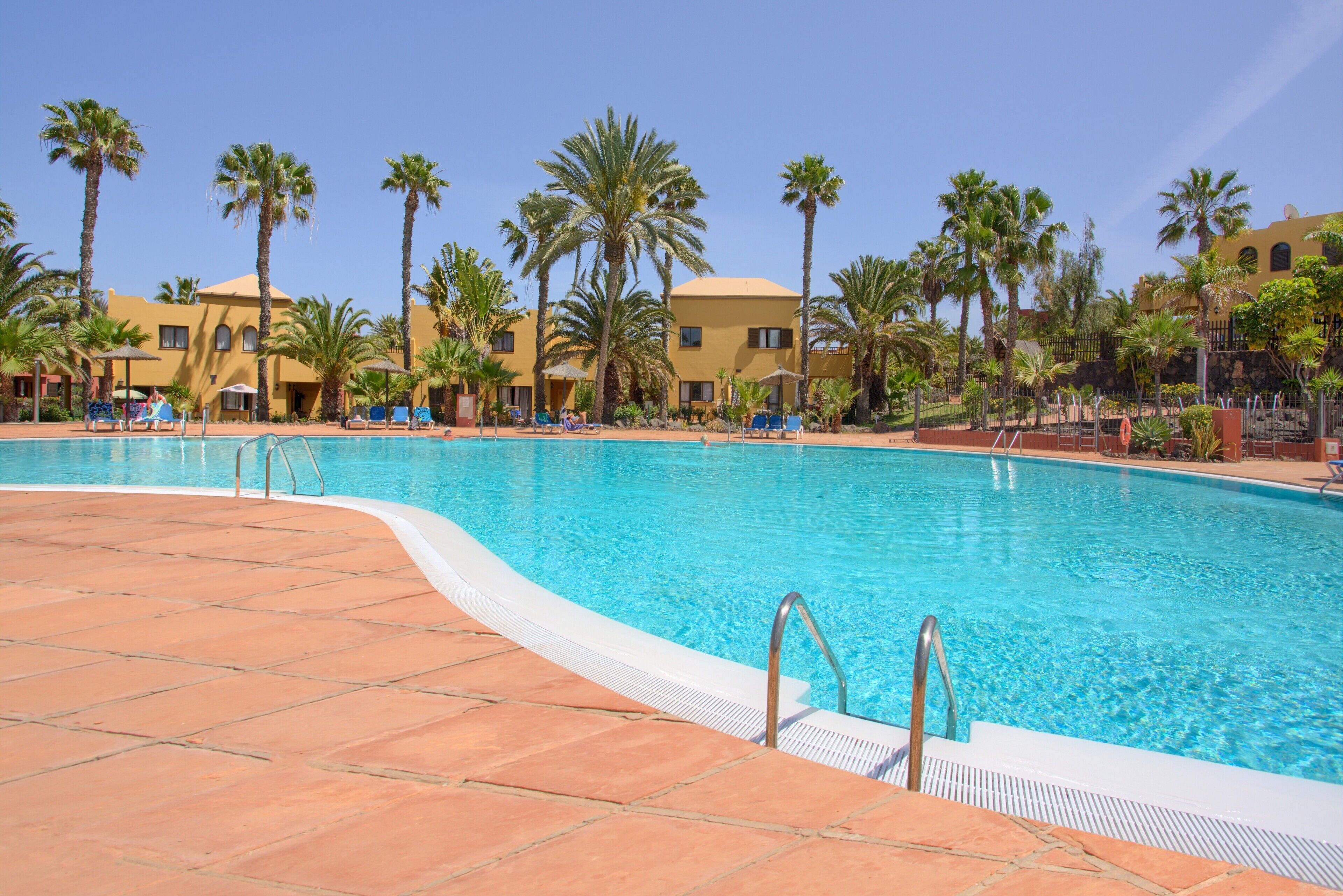 A swimming pool with clear blue water, surrounded by palm trees and yellow buildings.