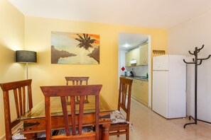 Un coin repas avec une table et des chaises en bois, un tableau au mur, une cuisine équipée d’armoires et d’un réfrigérateur, et un porte-manteau.