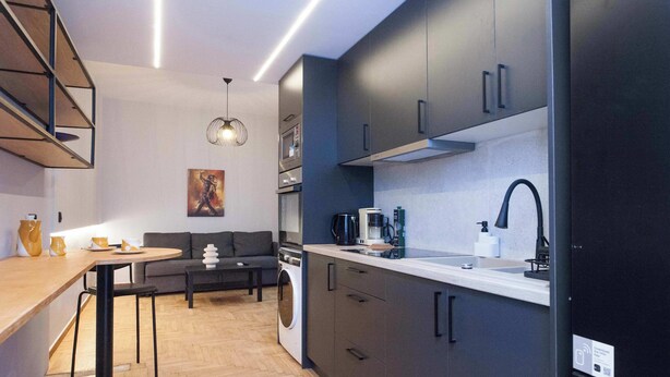 A modern kitchen with dark cabinets, a sink, and a microwave. There is a dining area with a table and chairs, and a sofa in the background. - Urban 2 Bd Home in Vibrant Galatsi (Galatsi)