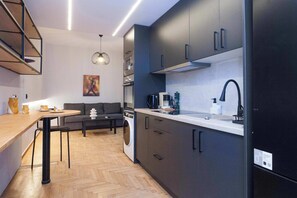 Une cuisine moderne avec des meubles de couleur sombre, un évier et un four à micro-ondes. On y trouve un coin repas avec une table et des chaises, ainsi qu’un canapé à l’arrière-plan.