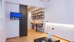A modern hotel room with a flat-screen TV, a small table with a vase, a chair, and a shelf with decorative items. - Urban 2 Bd Home in Vibrant Galatsi (Galatsi)