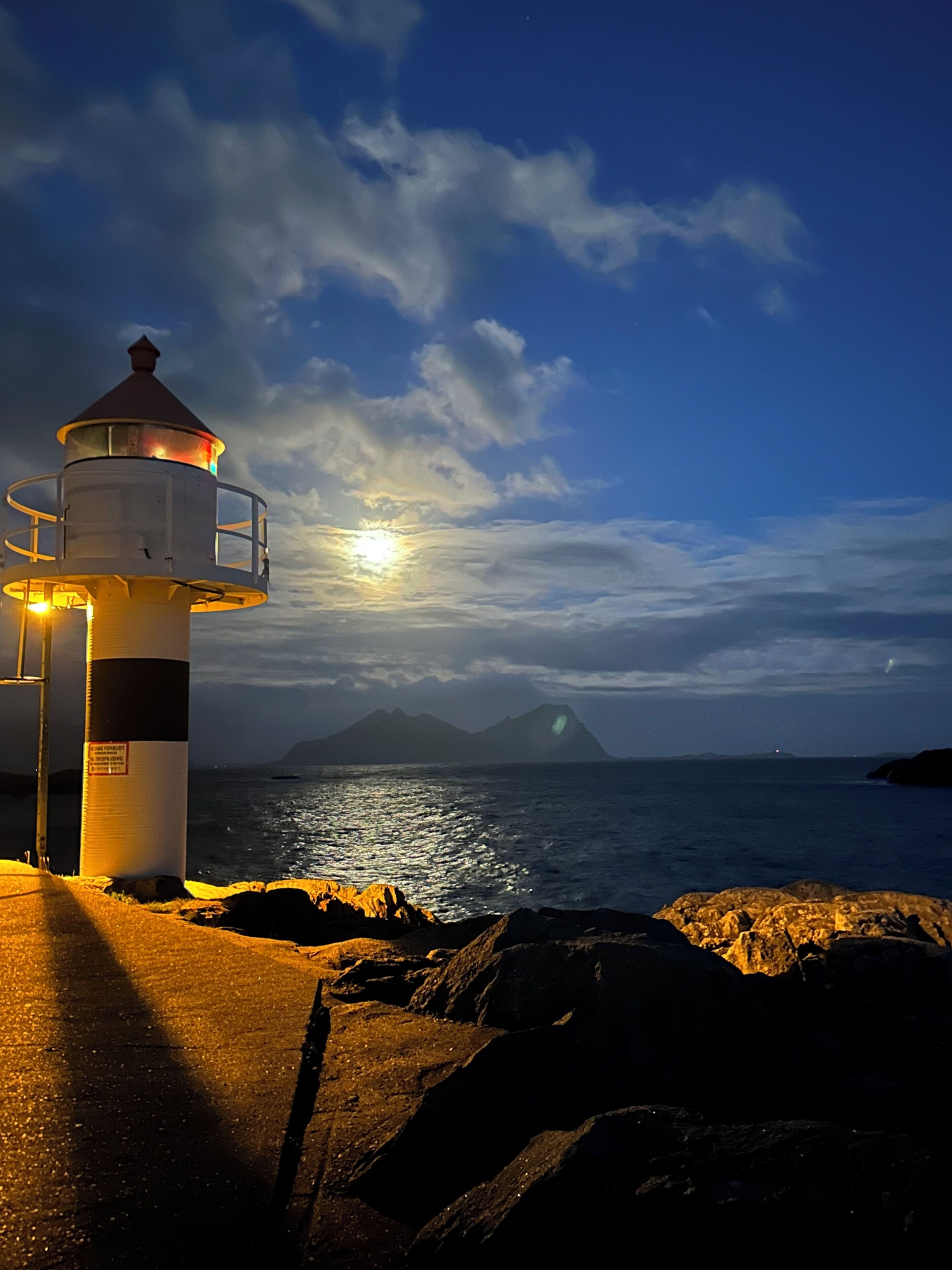 A lighthouse with a circular base and a conical top, situated on a rocky shoreline with a view of the sea and a mountain in the background.