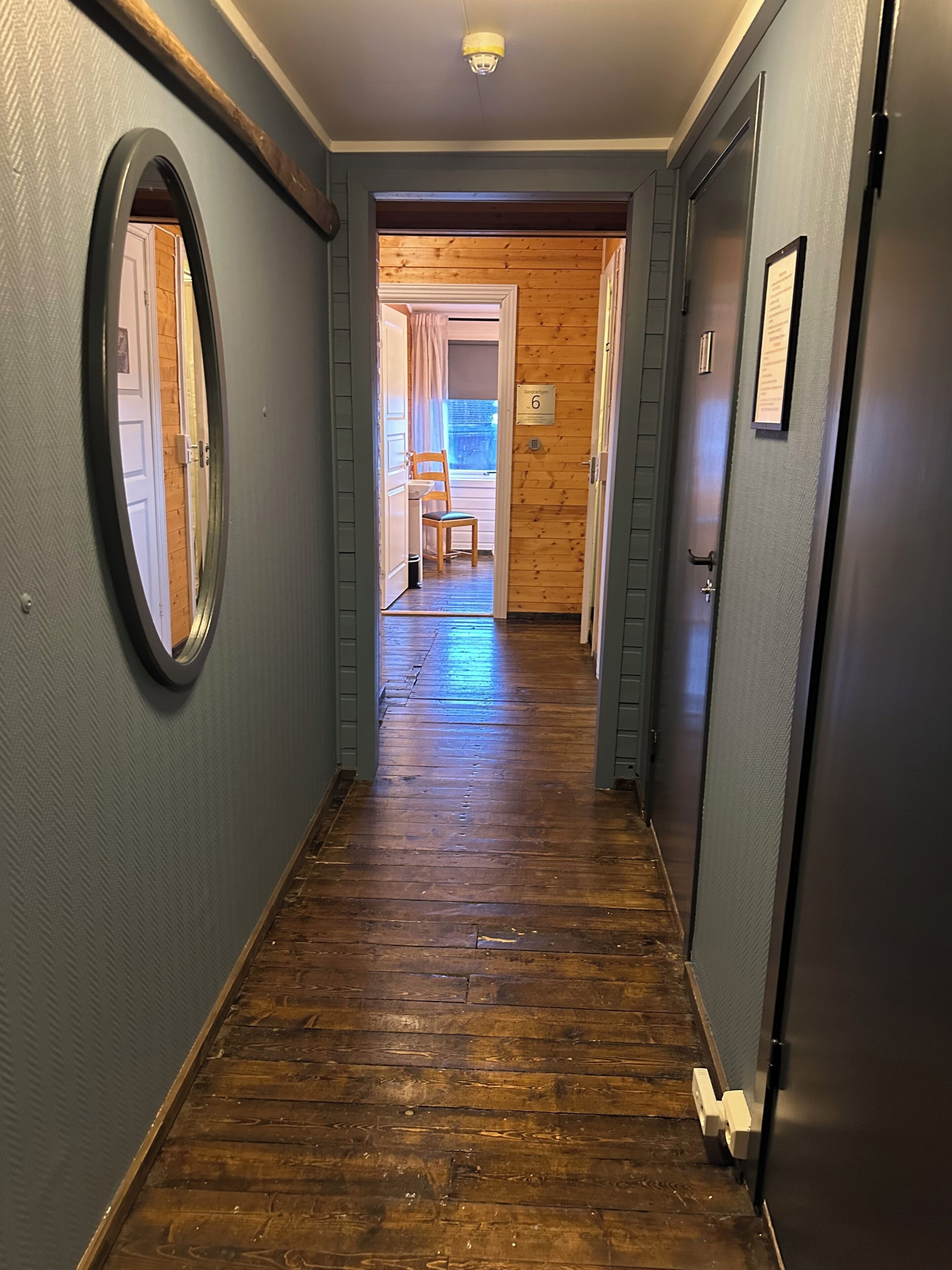 A hallway with wooden flooring, a mirror on the left wall, and a door on the right.