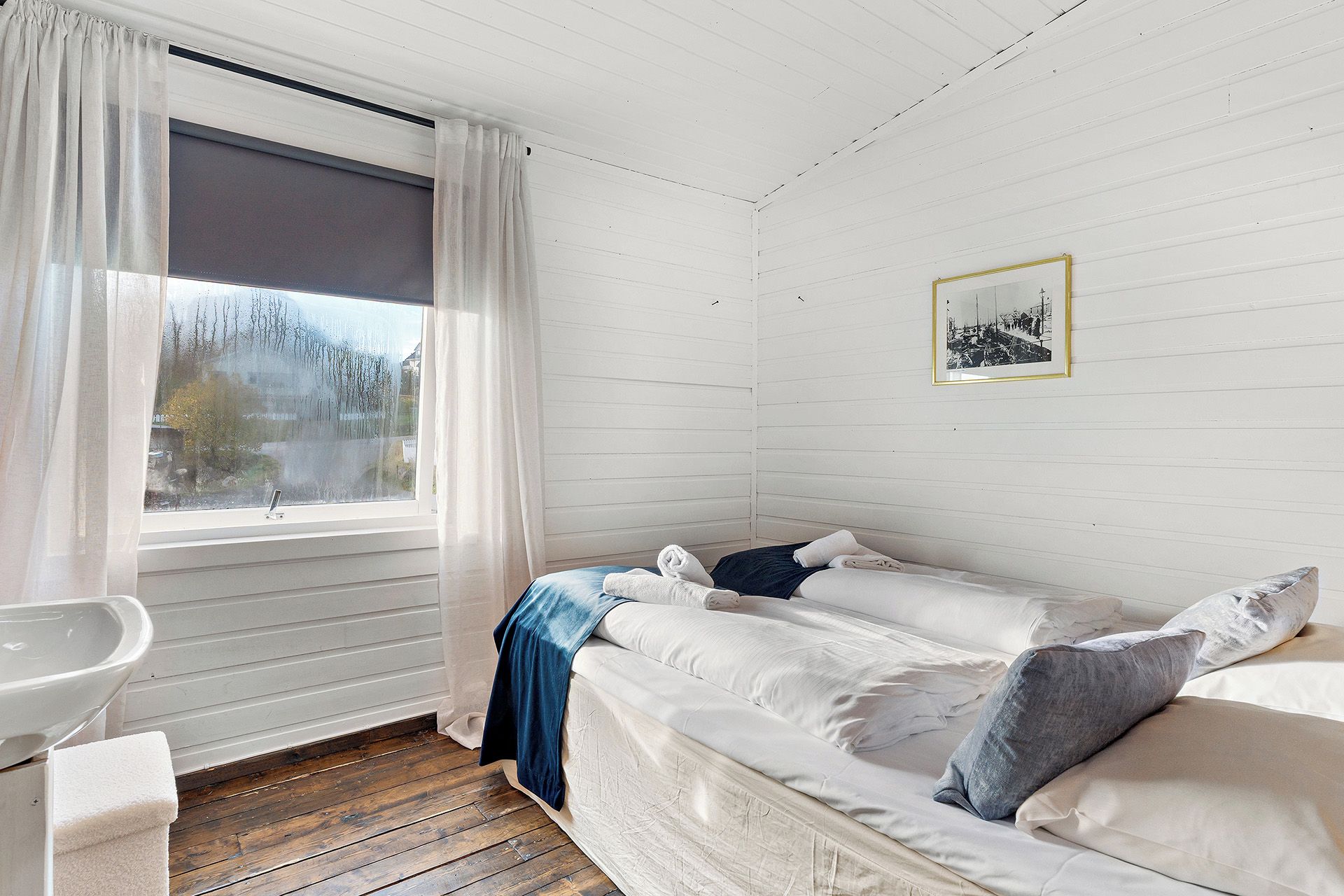 A room with a bed, a window with curtains, a sink, and a framed picture on the wall.