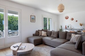 A modern living room with a sectional sofa, a round coffee table, and a dining area visible through glass doors.