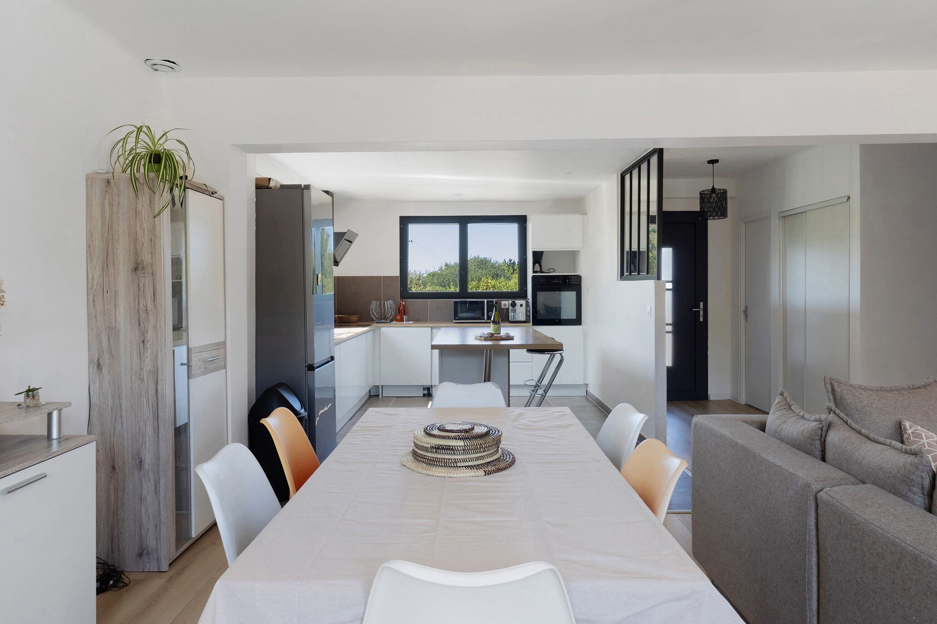 A modern kitchen with a dining area, featuring a long table, chairs, and a view through the window.