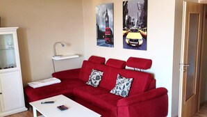 Ein Wohnzimmer mit einem roten Sofa, einem weißen Couchtisch und einem Schrank. Zwei an der Wand hängende Bilder zeigen Stadtansichten.