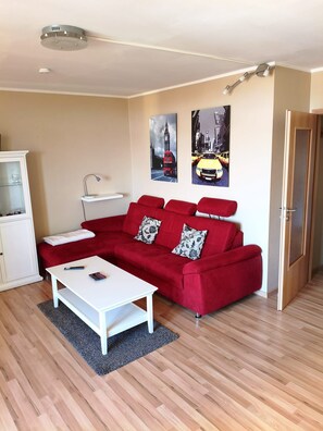 Ein Wohnzimmer mit einem roten Sofa, einem weißen Couchtisch und einem Schrank. Zwei an der Wand hängende Bilder zeigen Stadtansichten.