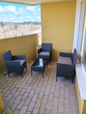 Ein Balkon mit Korbmöbeln und einem Glastisch, mit Blick auf ein bewaldetes Gebiet.