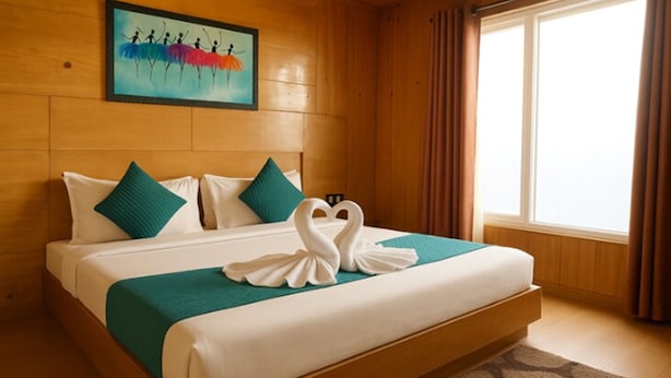 Habitación de hotel con cama, almohadas, una toalla con forma de corazón, un cuadro y una ventana con cortinas.