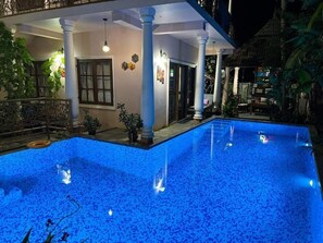 A swimming pool with a tiled deck, surrounded by a covered patio area with columns and potted plants.