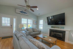 A living room with a sectional sofa, a fireplace, a television, and a ceiling fan. - OKI Hideaway: Ocean Views, Pool, Walk to Beach, Dog Friendly, Sleeps 7 (Oak Island)