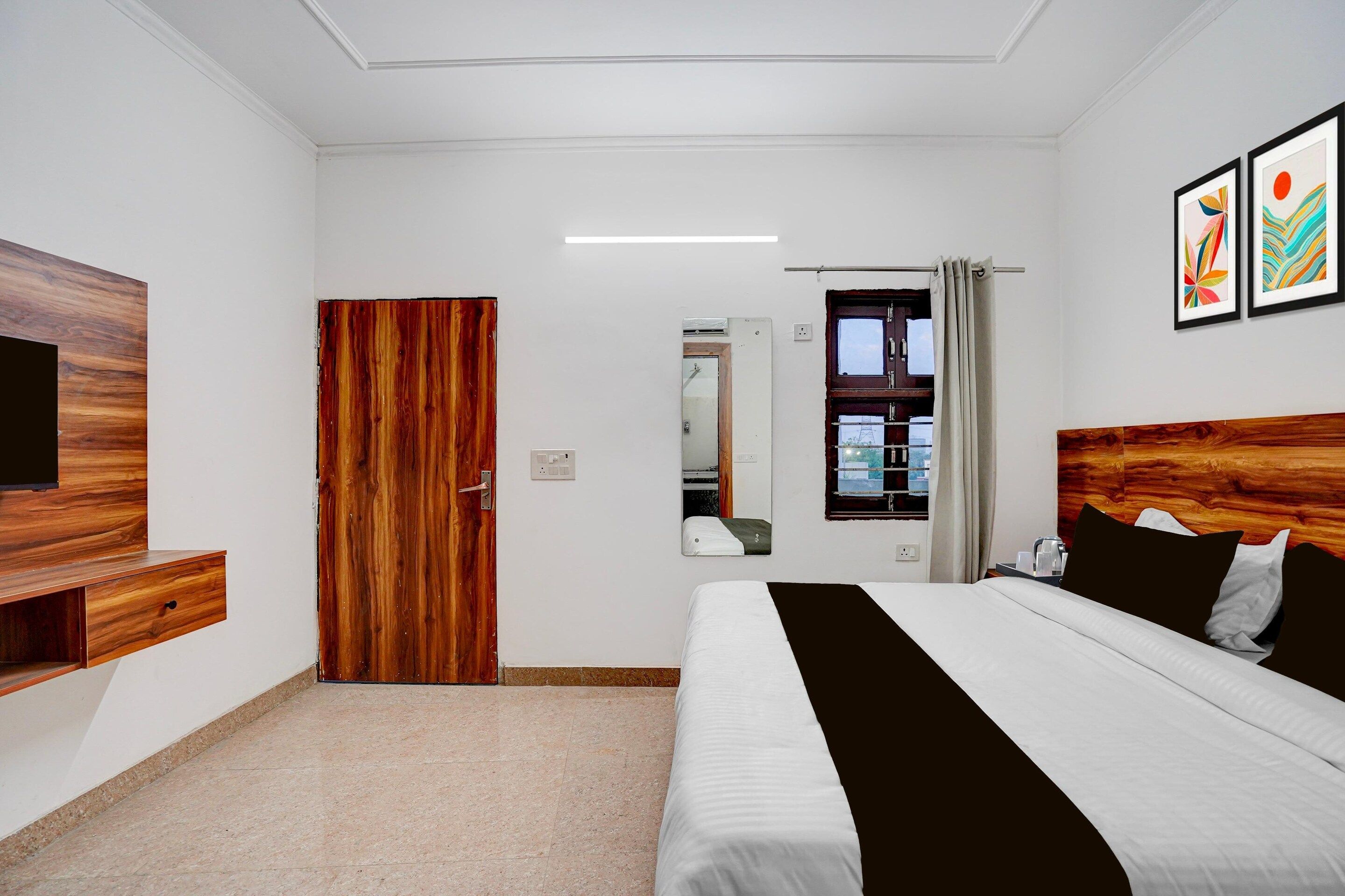 A hotel room with a bed, a TV, a wooden wardrobe, a mirror, and a window with curtains.