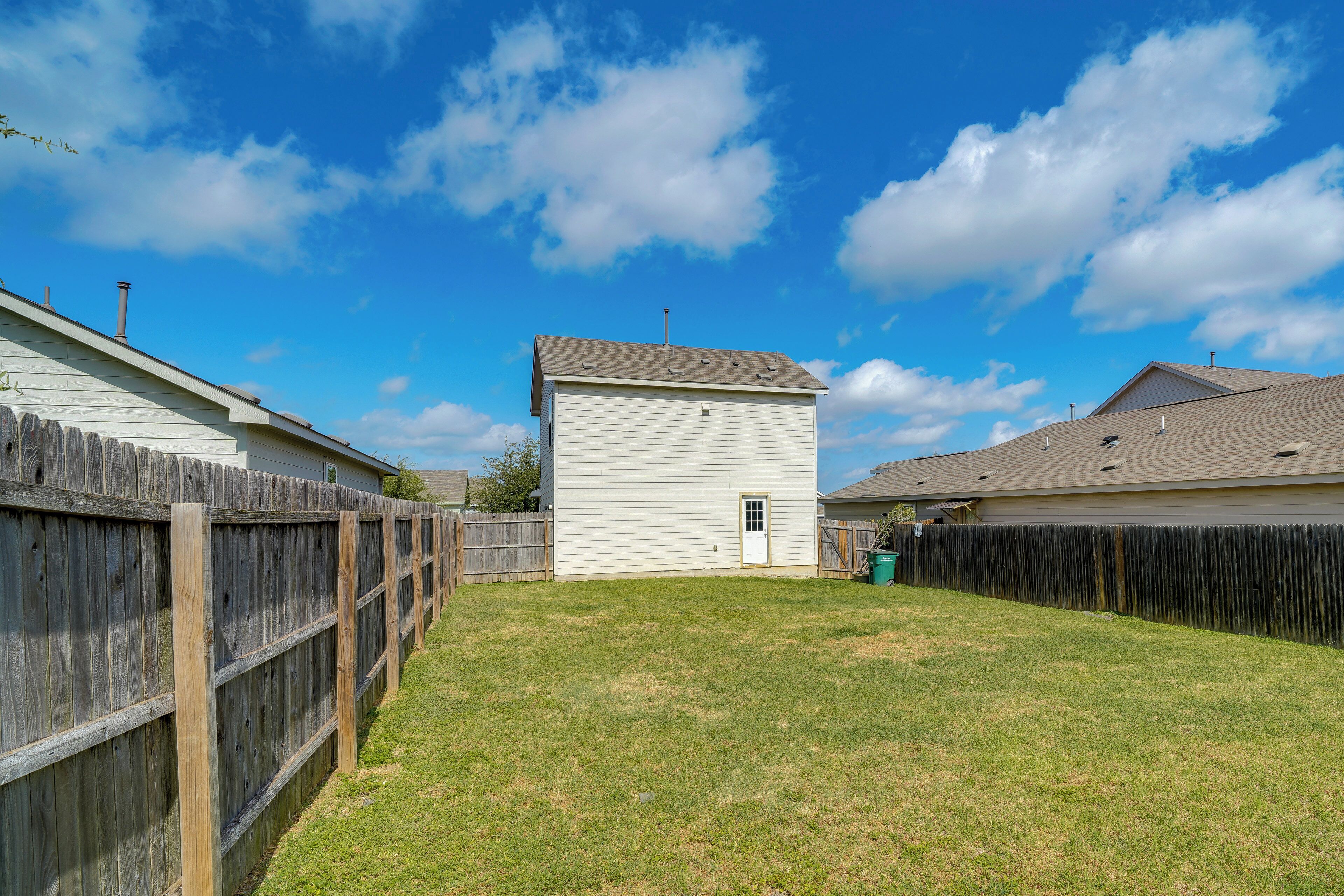 A fenced backyard with a white building, a wooden fence, and a clear blue sky.