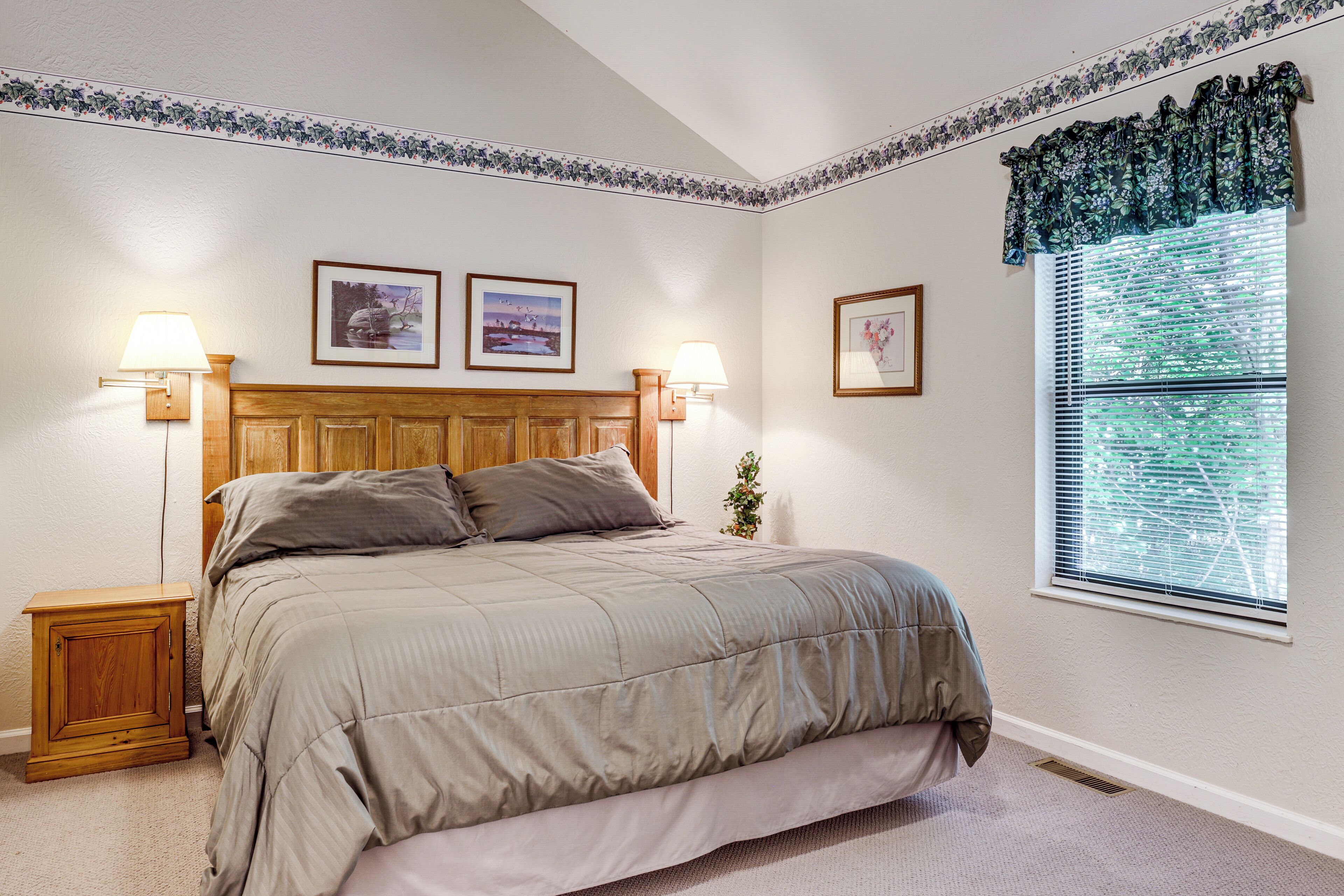 A bedroom with a bed, two wall-mounted lamps, a window with blinds, and framed pictures on the wall.