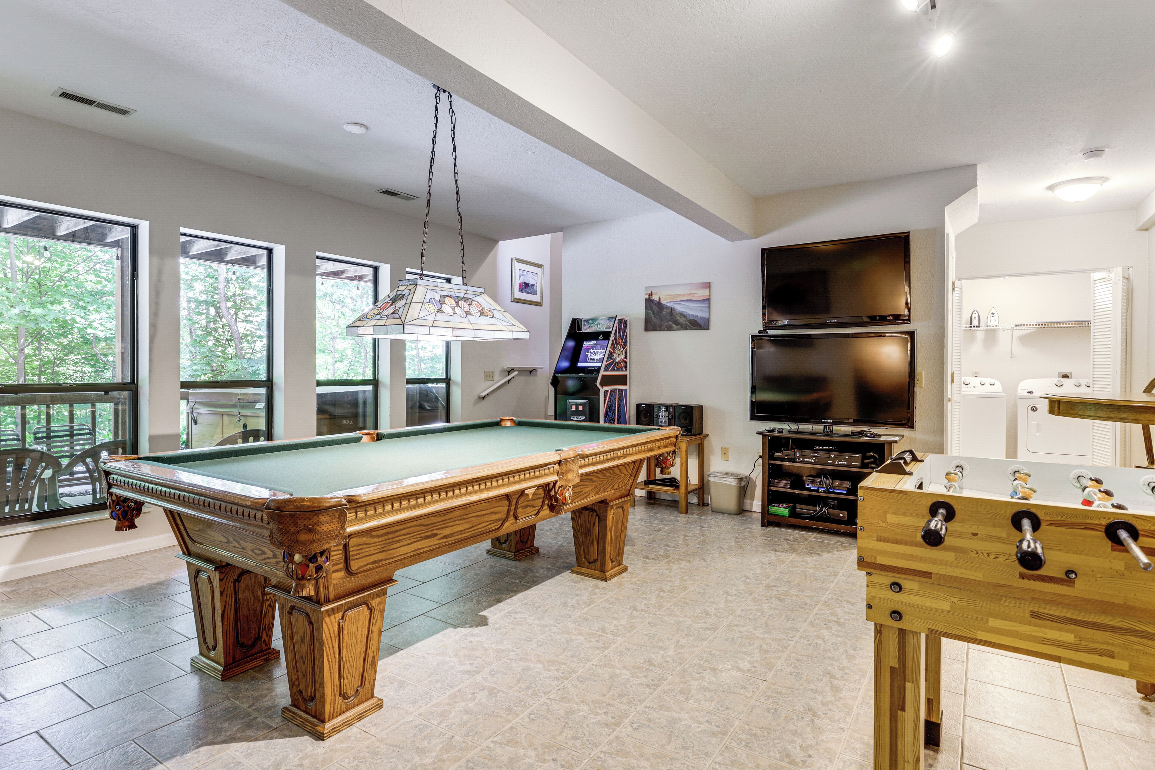 A pool table, a foosball table, and a television in a well-lit room with large windows.