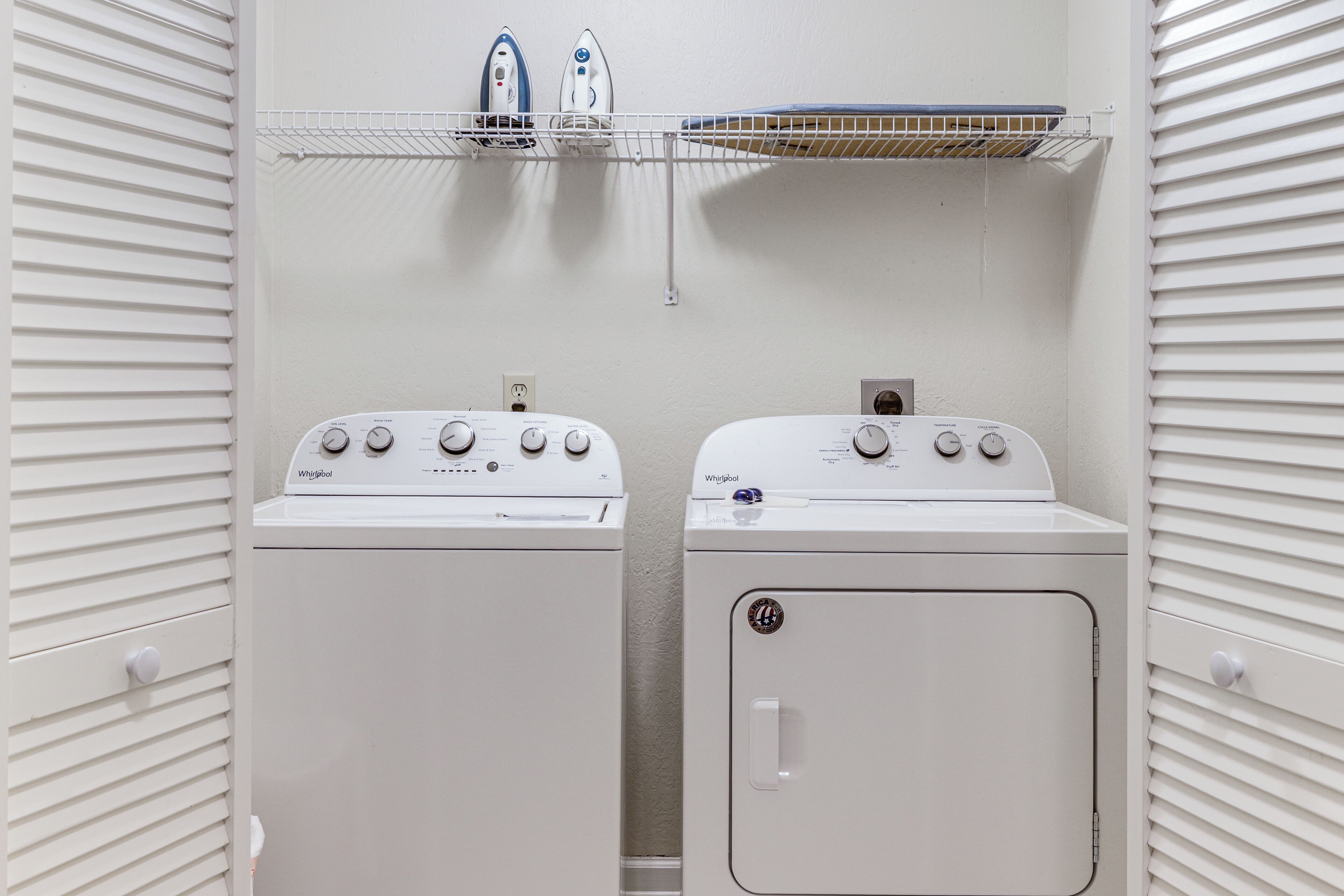 A laundry room with two Whirlpool washers and an iron on a shelf.