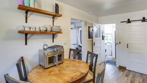 A dining area with a wooden table, chairs, a microwave, and shelves with kitchenware.