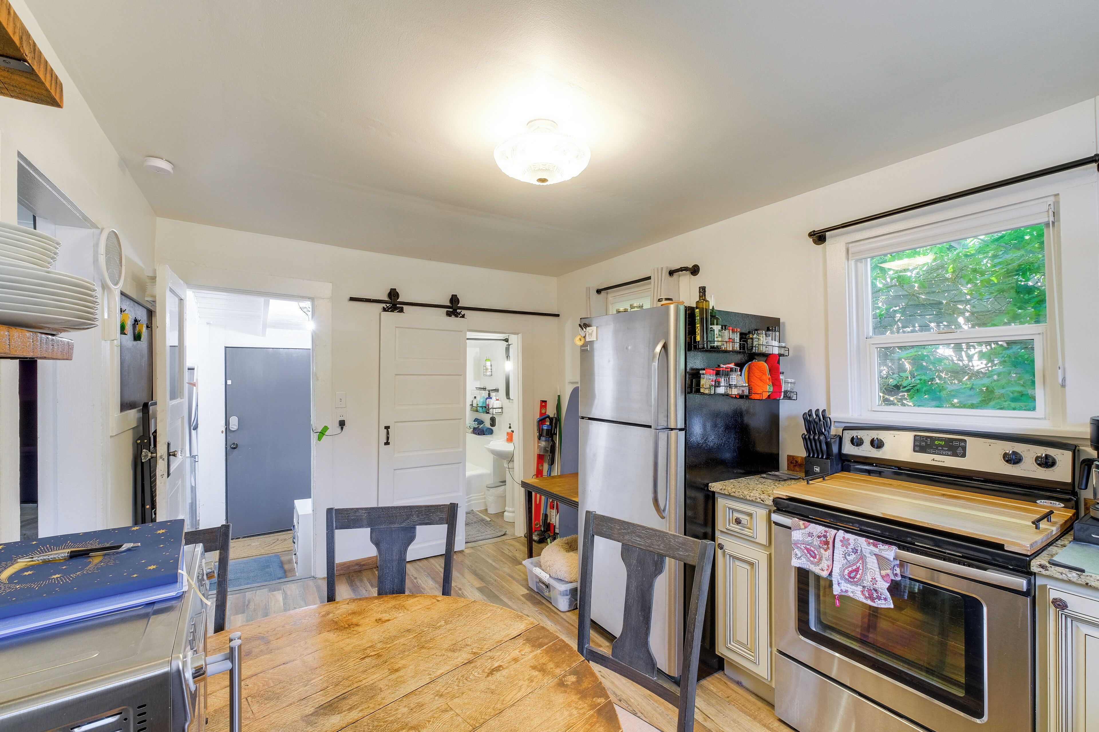 A kitchen with a wooden table, chairs, a refrigerator, a stove, and a window with a view of greenery.