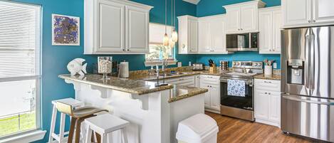 A kitchen with white cabinets, a stainless steel refrigerator, a granite countertop, and a window with blinds.