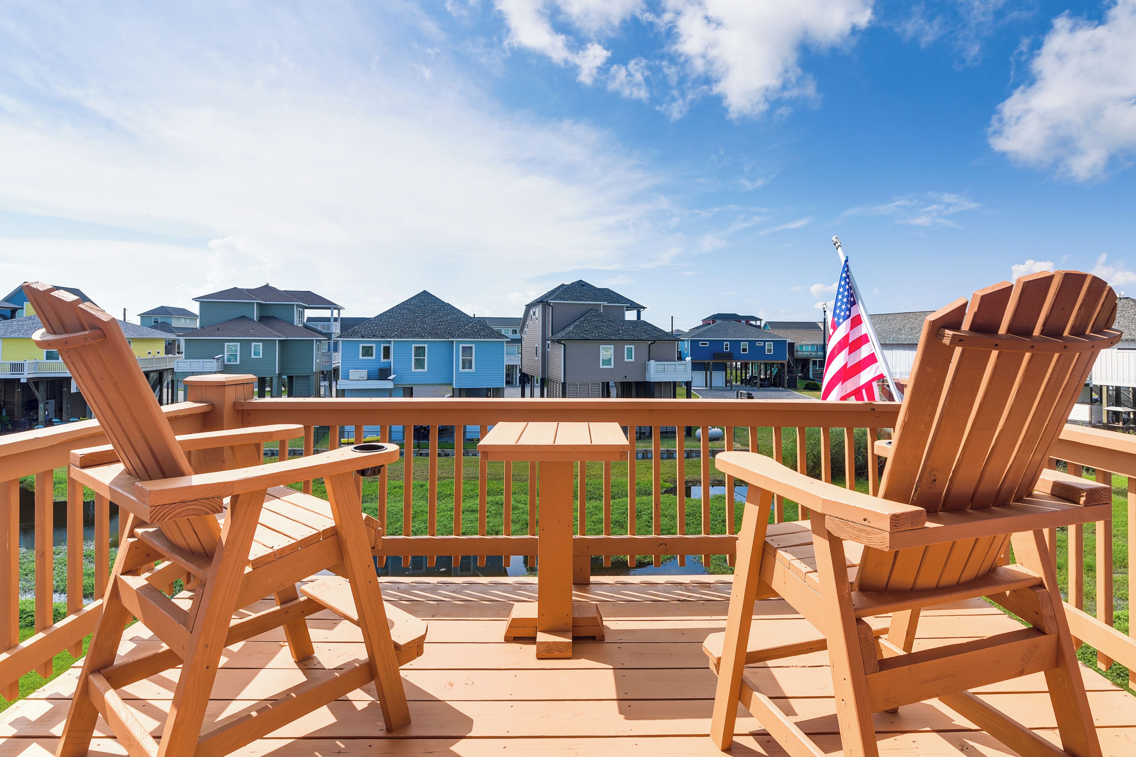 A wooden deck with Adirondack chairs and a small table, overlooking a row of colorful houses with an American flag.